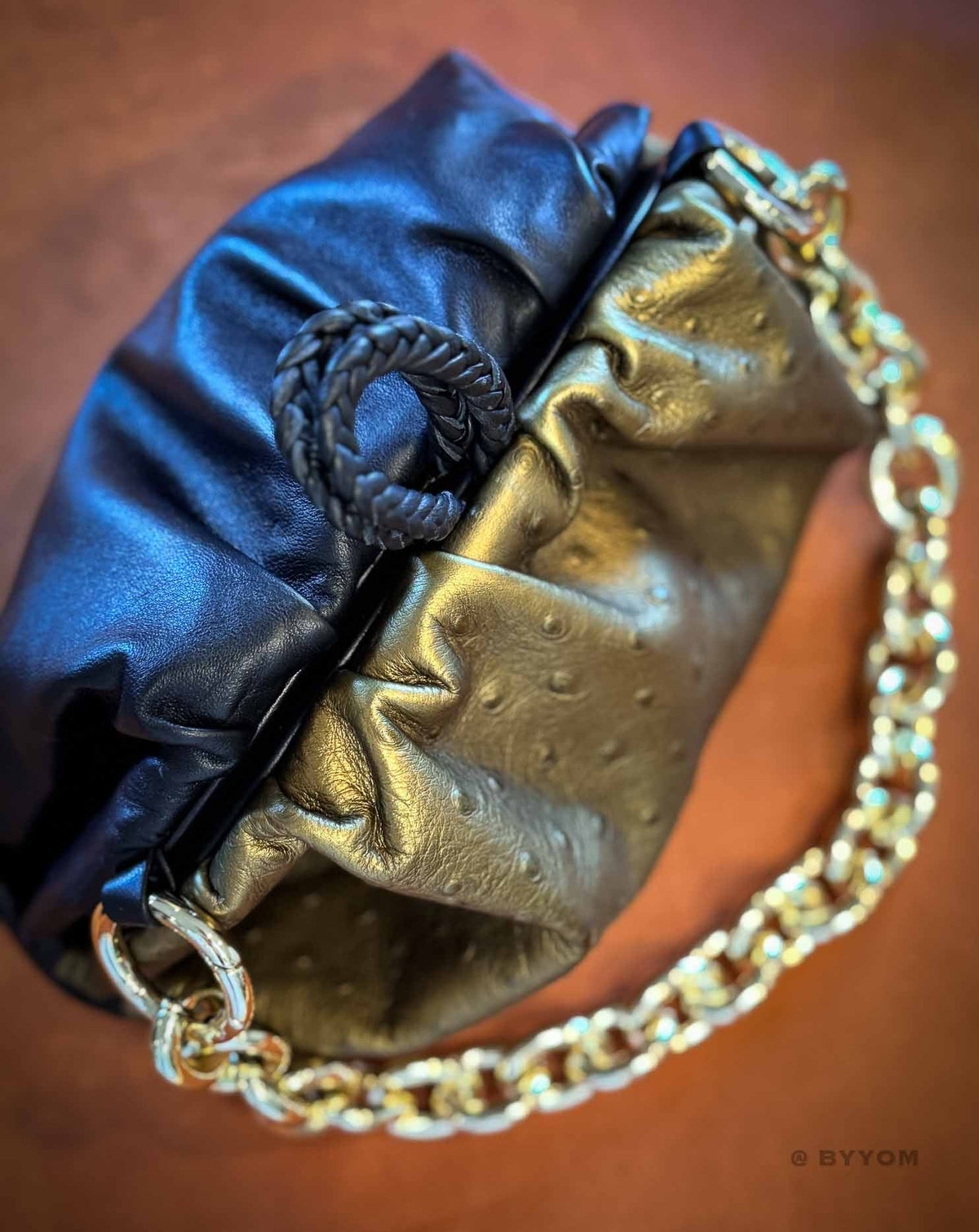 A close-up of a metallic clutch handbag with a gold ostrich-textured body, a dark blue satin flap, and a gold chain strap, set against a warm brown background.