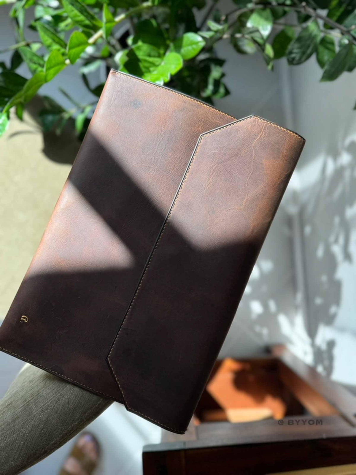 A brown leather folio or notebook cover held by a person's arm, with warm sunlight casting shadows across its surface. 