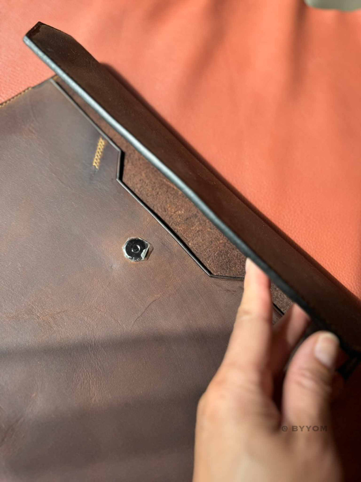 A close-up of a hand holding a dark brown leather tablet sleeve or folio case, showing the interior flap construction and a small metal lock or button. The background is a warm terracotta fabric.