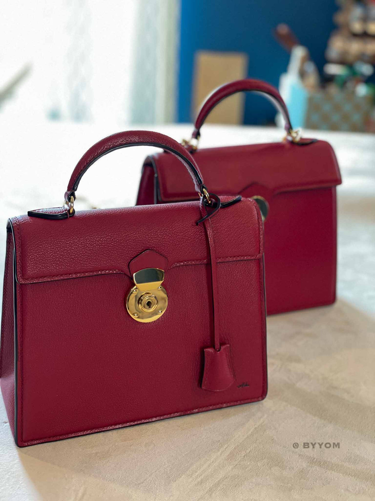 A structured red leather handbag with a top handle, gold-tone clasp, and hanging clochette, shown in front of a second matching bag on a light surface.